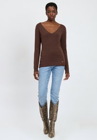 Brauner langärmeliger Pullover mit V-Ausschnitt, kombiniert mit hellblauen Jeans und gemusterten kniehohen Stiefeln mit Schlangenhautdesign.