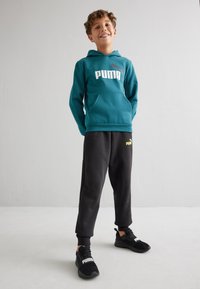 Sudadera con capucha en color azul verdoso con el logotipo blanco de "PUMA", combinada con pantalones de chándal negros que presentan un logotipo en color neón, y zapatillas negras. Textura de tela suave.