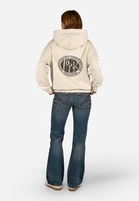 Sweatshirt bege com uma grande gráfica circular preta nas costas. Calças de ganga largas com um lavado azul e costuras detalhadas.