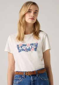 Balta medvilninė marškinėliai su mėlynu gėlių Levi's logotipu. Modelis dėvi rudą diržą su džinsinėmis aukšto liemens kelnėmis. Minkšta tekstūra, trumpomis rankovėmis.
