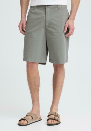 Mand iført grå knælange shorts og beige sandaler med dobbelt rem, vist fra talje til fødder mod en ensfarvet baggrund.