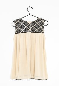 Mouwloos beige topje met een zwarte gemusterde schouderpartij, voorzien van een rond decolleté, knoopsluiting en een geplooide taille, hangend aan een zwarte haak.