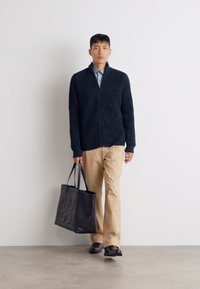 Cardigan de canalé oscuro, camisa de cuello azul claro, pantalones beige, zapatos de cuero negros y un gran bolso tote negro con un logo en relieve.