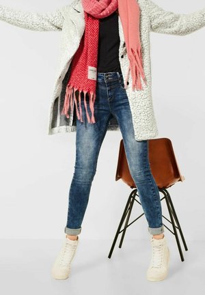 Person wearing white textured coat, red and pink scarf, blue jeans, and white sneakers standing with arms extended near a brown chair.