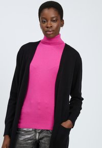 Zwarte cardigan over een felroze coltrui, gecombineerd met glanzende zilveren jeans. De cardigan heeft lange mouwen en een open voorkant.