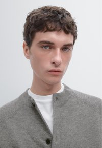 Cardigan en tricot gris avec un col montant et une fermeture à boutons, porté sur une chemise blanche. Il présente une texture douce et un design moderne et simple.