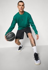 Grøn sweatshirt med sort logo, sorte shorts og sorte og hvide sportsko. Holder en sort basketball og viser ben-tatoveringer.