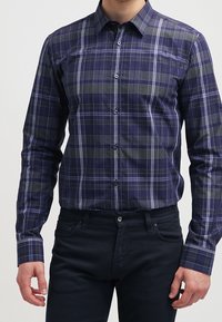 Camisa a rayas en tonos azul marino y verde con diseño de botones, mangas largas y cuello. Presenta líneas blancas contrastantes y un bolsillo en el pecho.