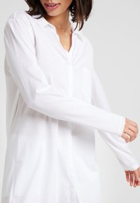 Camicia bianca a maniche lunghe realizzata in tessuto leggero, con chiusura a bottoni, colletto a punta e taschino sul petto. Texture liscia.