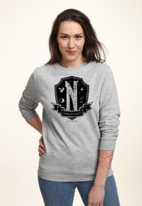 Mujer con cabello largo castaño vistiendo un sudadera gris con un emblema de escudo negro y el texto "Unitas Est Invicta", posando con una mano en la cadera.