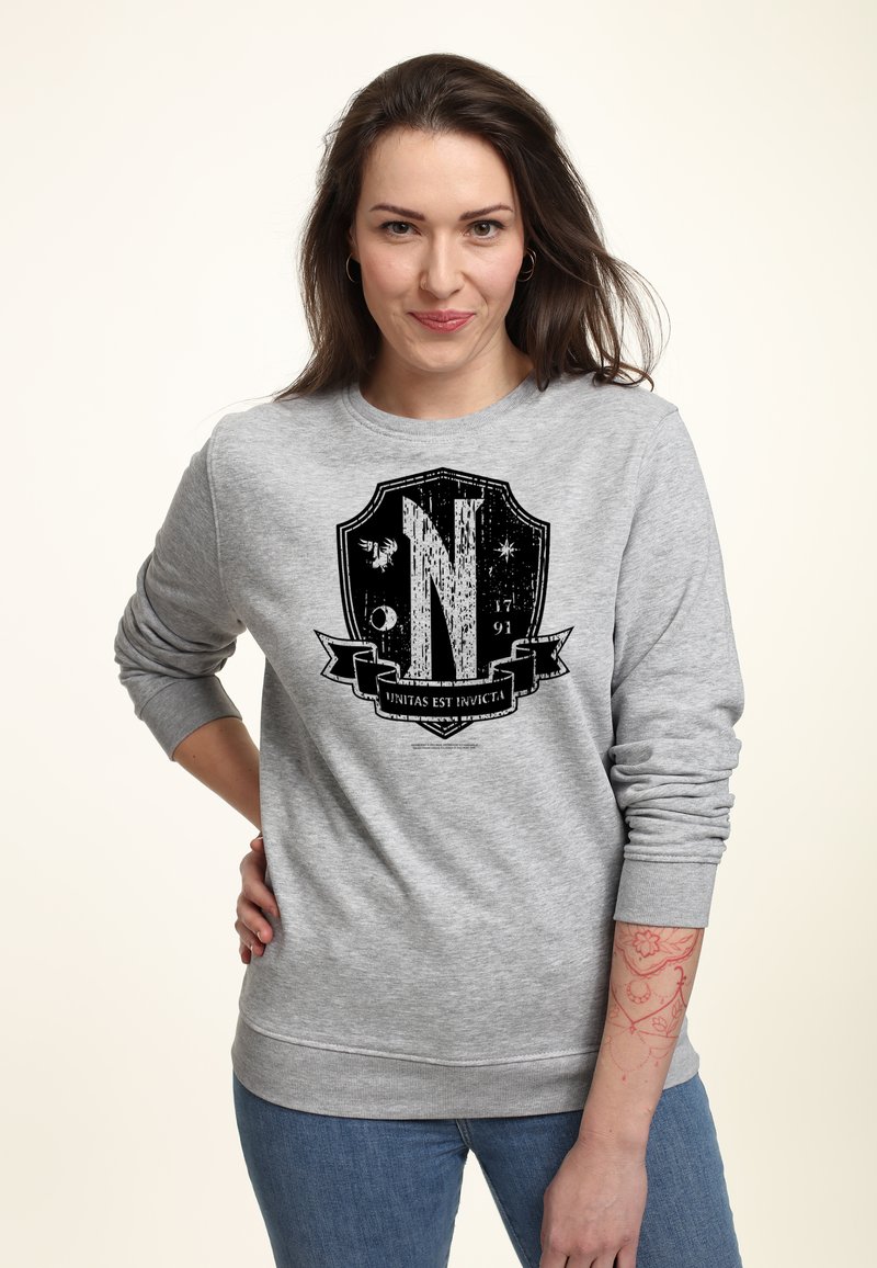 Mujer con cabello largo castaño vistiendo un sudadera gris con un emblema de escudo negro y el texto "Unitas Est Invicta", posando con una mano en la cadera.