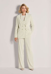 Vrouw in een crèmekleurige corduroy blazer met dubbele borst en wijde broek, staand tegen een lichtbeige achtergrond.