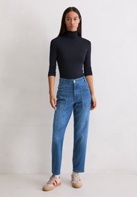 Jeune femme debout portant un col roulé noir, un jean taille haute bleu avec de grandes poches avant, et des baskets beiges avec des rayures rouges.