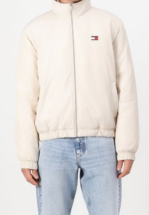 Man draagt een lichtbeige jas met ritssluiting en elastische manchetten, gecombineerd met lichtblauwe spijkerbroek, staand tegen een witte achtergrond.