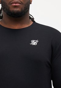 Black long-sleeve shirt made of cotton with a round neck, featuring a white embroidered logo on the upper left side.