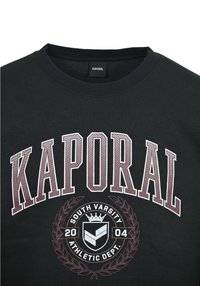 Sudadera negra de manga larga con el texto "KAPORAL" en letras granate contorneadas. Incluye un emblema gráfico y un diseño de corona de olivo.