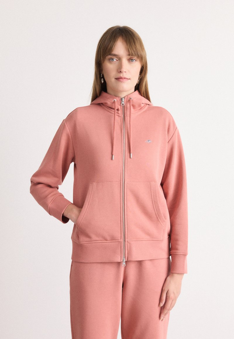 Roza zip-up dukserica izrađena od mekog materijala, sa prednjim džepom, vezicama i rebrastim manžetama. Jednostavna oznaka na grudima.