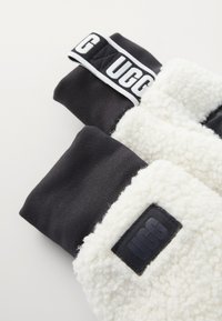 Juodi ir balti UGG pirštinės su pūkiniais rankogaliais ir prekės ženklo audinio bei odiniais logotipais ant šviesaus fono.