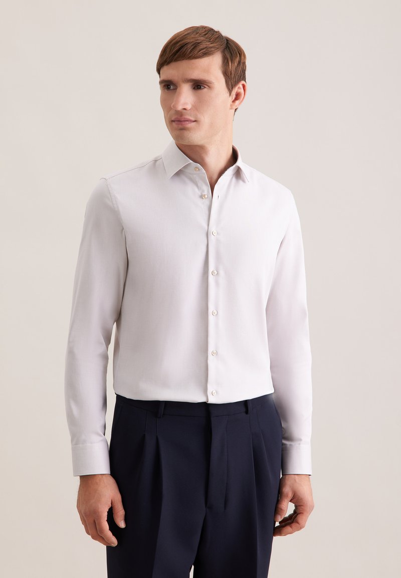 Chemise gris clair à manches longues avec une texture douce, col boutonné et légère brillance, associée à un pantalon bleu marine foncé.