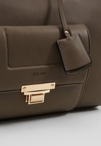 Gros plan d’un sac à main en cuir marron avec une poche à rabat sur le devant, une fermeture dorée, et une petite étiquette en cuir attachée, marquée du logo "PICARD".