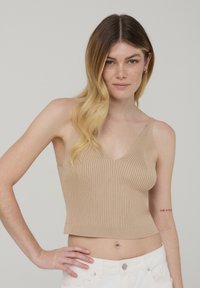 Top in maglia a coste beige con scollatura a V e design cropped. Dotato di spalline sottili e texture liscia, indossato con pantaloni bianchi.