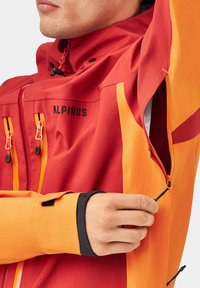 Czerwona i pomarańczowa wodoodporna kurtka z zamkami błyskawicznymi, elastycznymi mankietami na nadgarstki i logo. Posiada design z mieszanymi kolorami oraz regulowane detale.