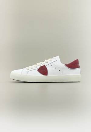 Zapatilla de cuero blanco con cordones beige, talón y parches laterales de ante rojo, exhibida sobre un fondo gris neutro, vista lateral.