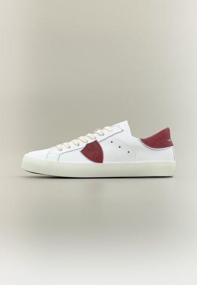 Baskets en cuir blanc avec lacets beiges, talon en daim rouge et patchs latéraux, présentées sur un fond gris neutre, vue de profil.