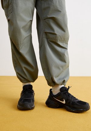 Persoon die losse groene broek en witte sokken met zwarte Nike-sneakers draagt, staand op een gele ondergrond tegen een lichte achtergrond.
