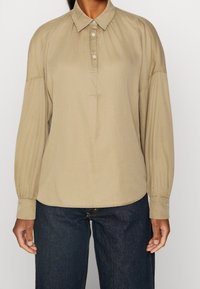 Chemise beige à manches longues avec col à boutons, présentant des épaules plissées et des poignets à boutons. Tissu lisse avec une coupe décontractée.