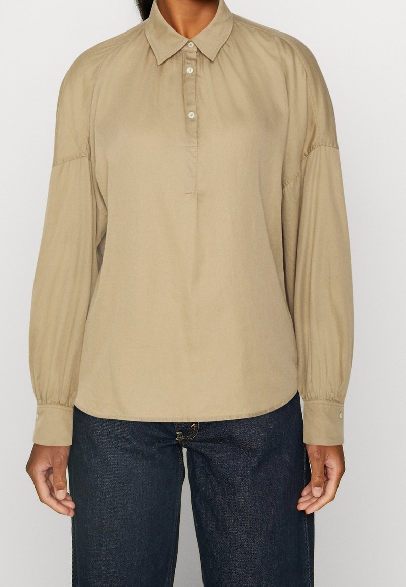 Chemise beige à manches longues avec col à boutons, présentant des épaules plissées et des poignets à boutons. Tissu lisse avec une coupe décontractée.