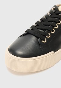 Black leather sneaker with a rounded toe, textured upper, gold eyelets, black laces, and a ribbed cream-colored sole.
