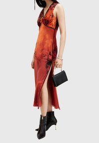 Rotes Blumen-Kleid mit V-Ausschnitt, taillierten Design, seitlichem Schlitz und schwarzen Akzenten. Kombiniert mit einer kleinen schwarzen Handtasche und Stiefeletten.