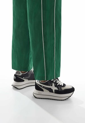 Sneaker neri e grigi con una spessa suola bianca e dettagli argento. Presentano un design a stella e sono abbinati a pantaloni larghi verdi con strisce bianche.