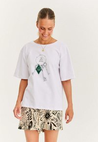 Uśmiechnięta kobieta z włosami związanymi w kok, ubrana w białą, oversize'ową koszulkę z grafiką klucza oraz wzorzyste beżowe szorty, na tle jednolitego tła.