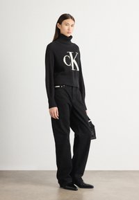 Maglione nero a collo alto con texture a coste e logo "CK" bianco, abbinato a pantaloni larghi neri e stivaletti, mentre tiene una borsa piccola.