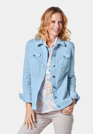 Veste bleu clair à boutons, en tissu lisse, avec deux poches poitrine, des manches retroussées et une coupe droite. Portée par-dessus une chemise à motifs.