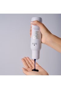 Bottiglia di shampoo bianca con testo dorato, che versa un liquido blu scuro in una mano. Texture liscia e lucida, forma cilindrica e beccuccio affusolato.