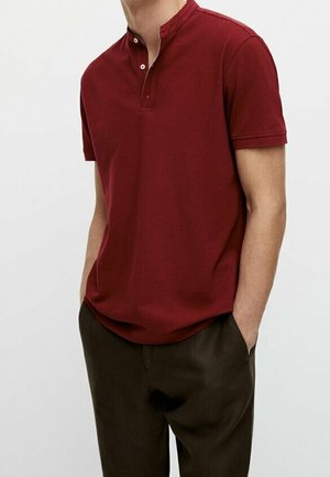 Mand iført en kortærmet burgunder henley-shirt løst stoppet ned i mørkebrune bukser med hænderne i lommerne, stående foran en ensfarvet baggrund.