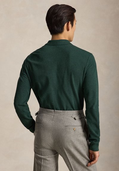 Man met een donkergroen overhemd met lange mouwen en lichtgrijze broek, met de rug naar een neutrale achtergrond.