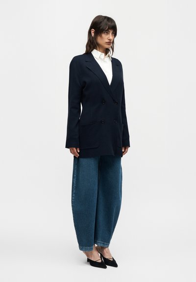 Γυναίκα που στέκεται φορώντας λευκό πουκάμισο, υπερμέγεθος σκούρο μπλε σακάκι, φαρδιά μπλε τζιν και μαύρα χαμηλά παπούτσια με μυτερή μύτη σε λευκό φόντο.