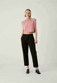 Woman standing wearing a sleeveless pink crop top with padded shoulders, black high-waisted trousers, and black heeled mules.