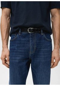 Jeans azul oscuro con un ajuste clásico, que presentan un diseño de bolsillo con costuras, usados con un cinturón de cuero negro y una camiseta azul marino.