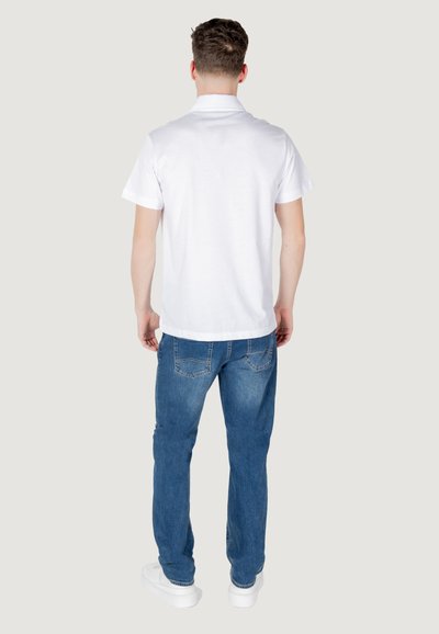 Polo blanc à manches courtes, associé à un jean en denim bleu. Le modèle se tient de dos face à l'appareil photo, portant des baskets blanches.