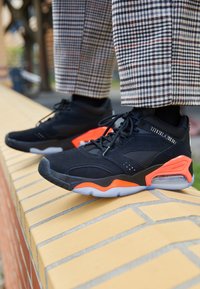 Scarpe sportive nere con dettagli arancioni e un design testurizzato, dotate di colletto imbottito e chiusura con lacci, posizionate su una sporgenza di mattoni.