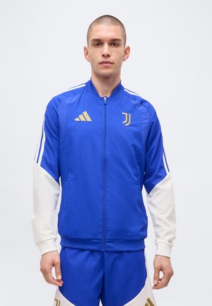 Homme portant un survêtement Adidas Juventus bleu et blanc avec des logos dorés, debout devant un fond uni clair.