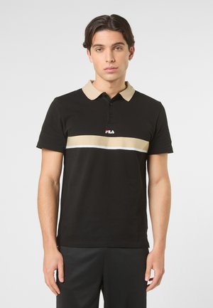 Młody mężczyzna o krótkich ciemnych włosach, ubrany w czarną koszulkę polo z beżowym kołnierzykiem i poziomymi beżowo-białymi paskami na wysokości klatki piersiowej.