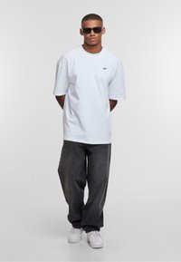 Camiseta blanca de gran tamaño con logotipo bordado, combinada con jeans loose fit en gris oscuro y zapatillas blancas. El modelo lleva gafas de sol oscuras.