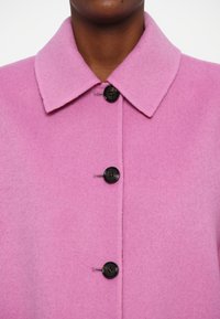 Palton roz din lână cu guler și patru nasturi negri. Țesătura are o textură moale, prezentând un finisaj neted și un design minimalist.