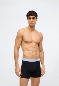 Svarta bomullsboxershorts med en grå midjeresår med Calvin Klein-logotyp, med en åtsittande design och slät textur. Modellen står med armarna avslappnade.